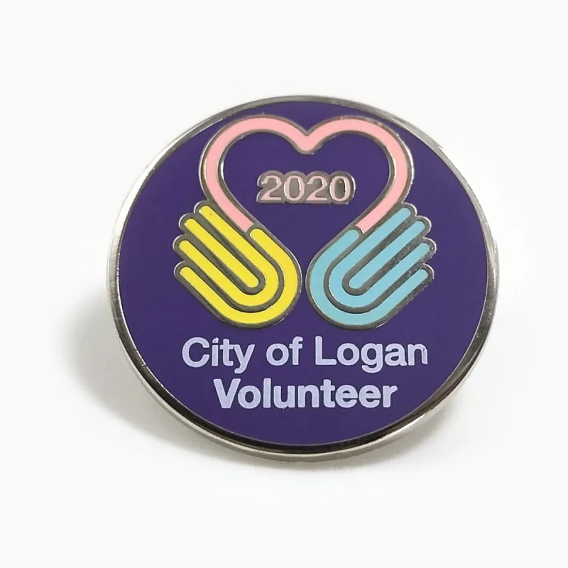Pin redondo de esmalte duro sobre fondo morado con dos manos que forman un corazón, el año 2020 y el texto «Volunteer».