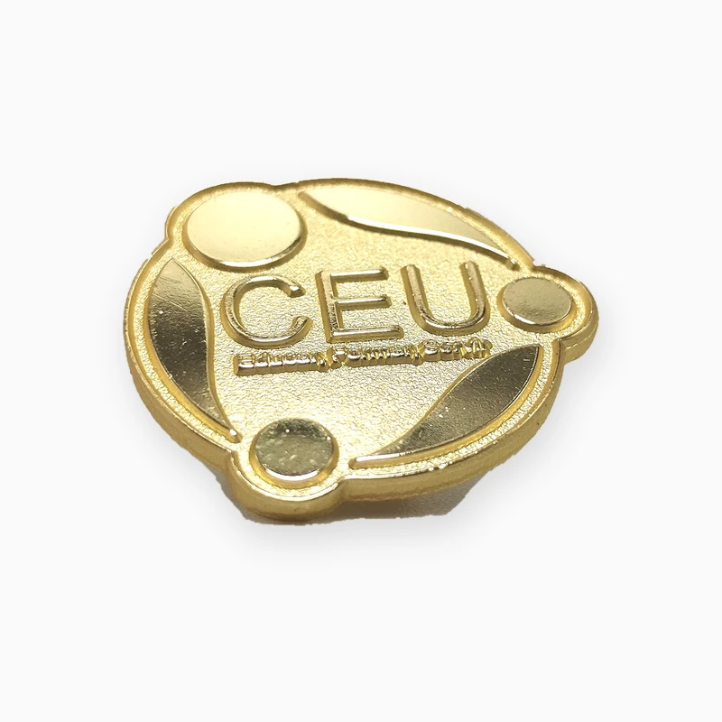 Pin metálico troquelado dorado con el logotipo CEU en relieve, círculos pulidos y fondo texturizado arenado, sin esmalte.