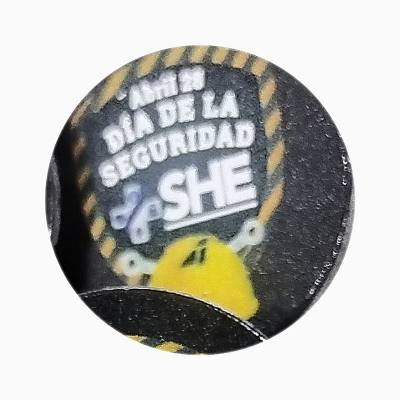 Pin redondo negro con impresión UV de franjas amarillas de advertencia, un casco y un texto sobre el día de la seguridad
