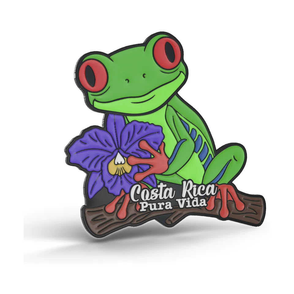 Imán de metal recortado de una rana de ojos rojos sobre una rama junto a una orquídea morada, con el texto Costa Rica Pura Vida.